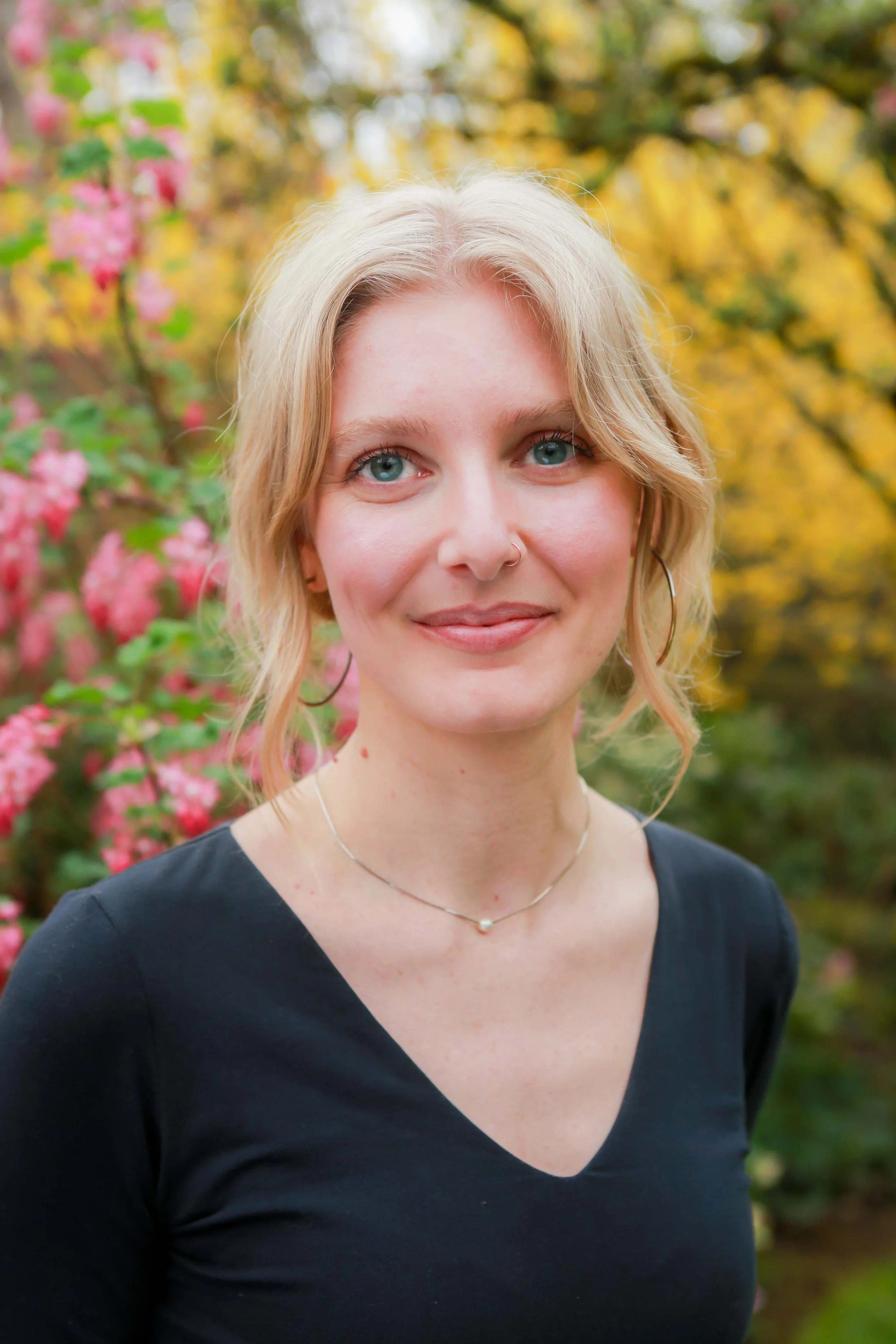 A young woman with blonde hair and blue eyes, wearing a black V-neck shirt, a delicate necklace, and hoop earrings, posing outdoors with pink flowers and yellow autumn leaves in the background.