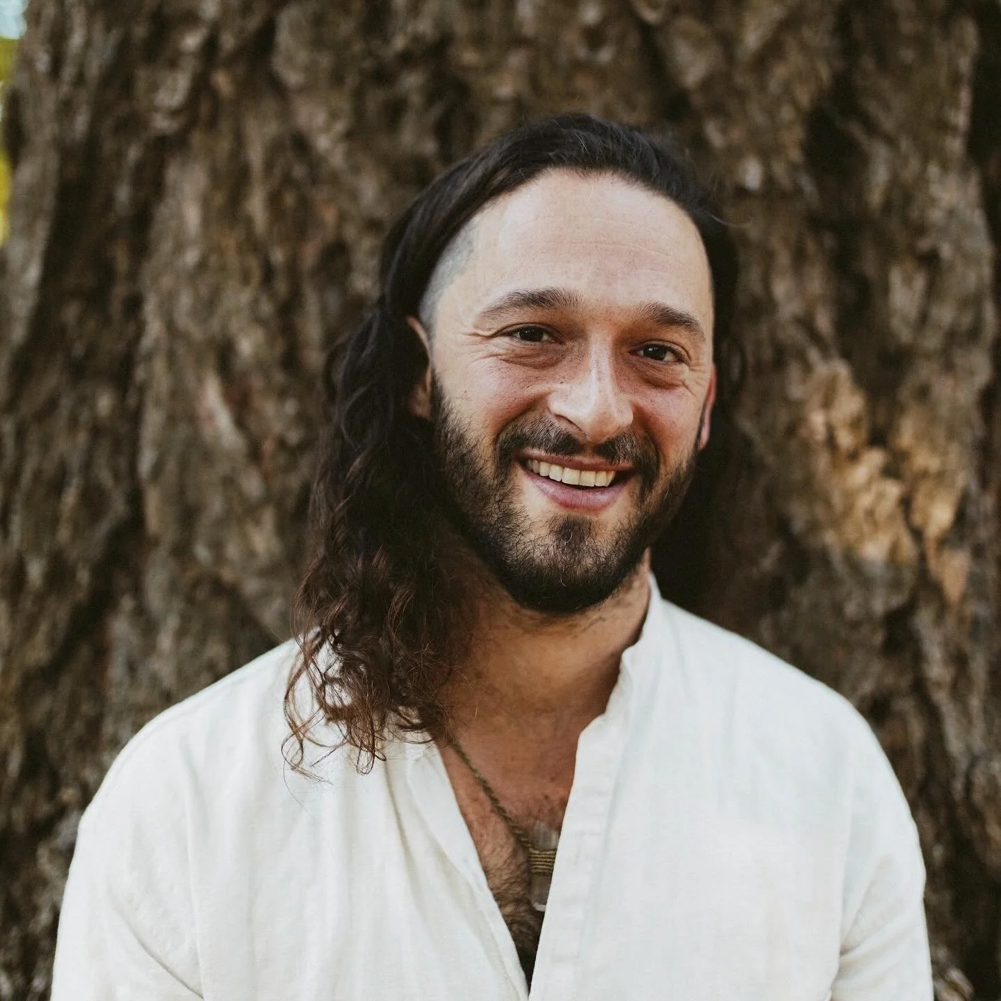 A smiling man with long dark hair and a beard, wearing a white shirt, standing in front of a large tree trunk.