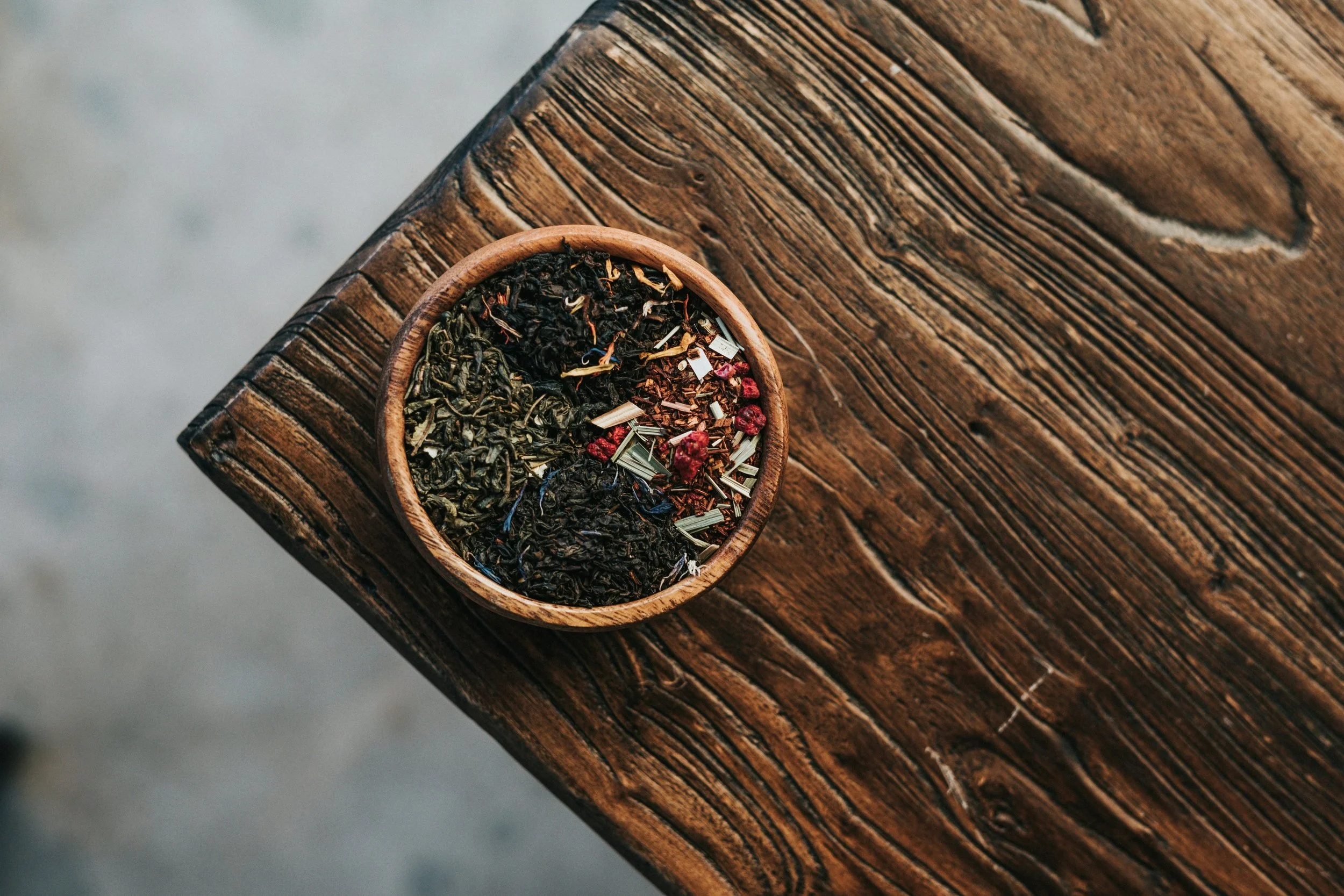 A round wooden bowl filled with loose herbal tea leaves, including dried flowers and herbs, placed on a rustic wooden table.