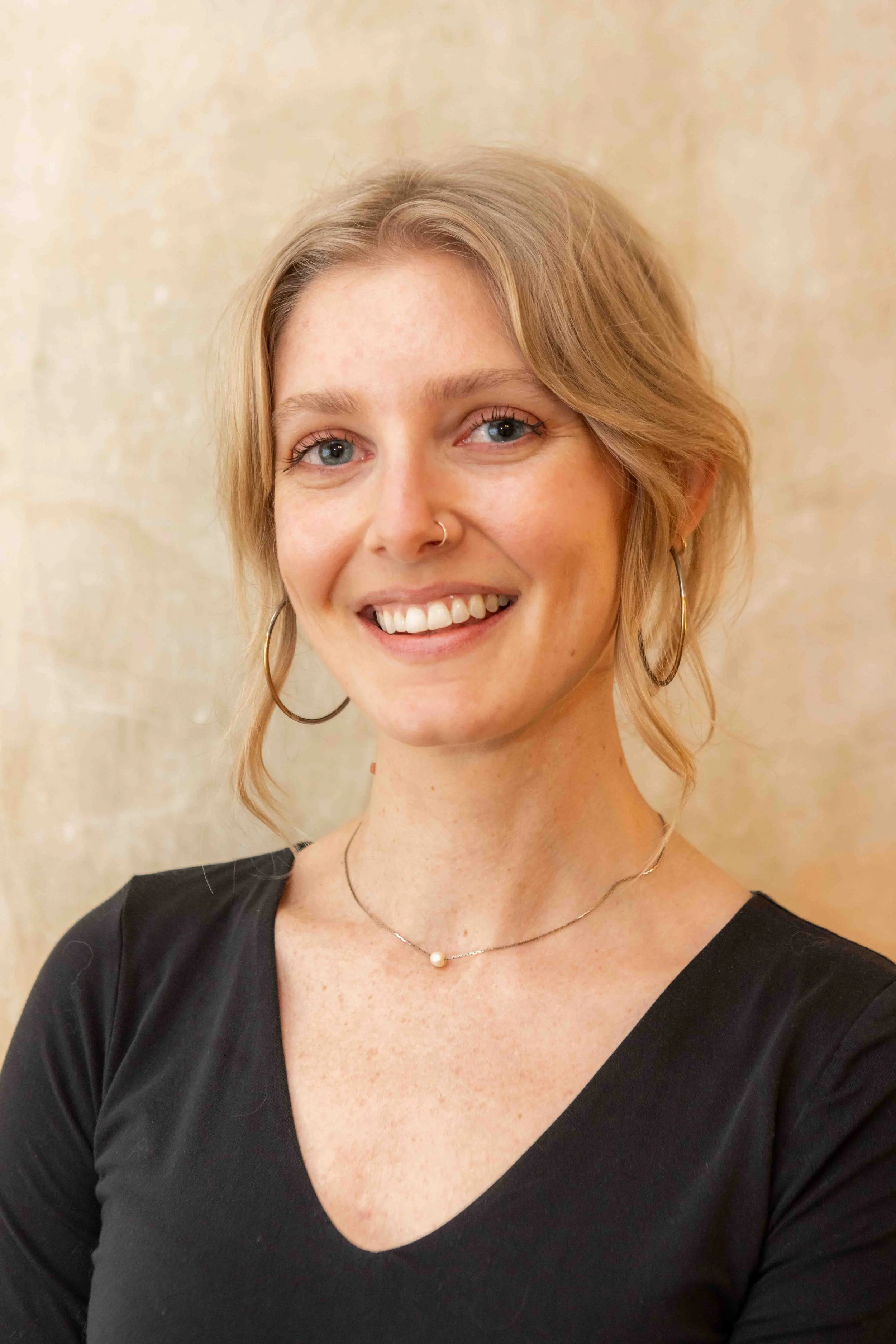 Close-up of a young woman with blonde hair, blue eyes, wearing hoop earrings, a small nose ring, a necklace with a pearl, and a black top, smiling against a beige textured background.