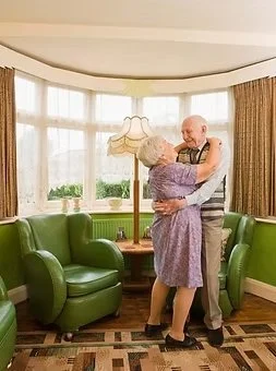 An elderly couple dances happily in a cozy, well-lit senior living space at Cedars Assisted Living, surrounded by elegant green armchairs and large windows that create a warm and inviting atmosphere