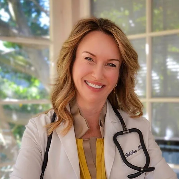 Smiling female physician in white coat with stethoscope, sitting by a large window.