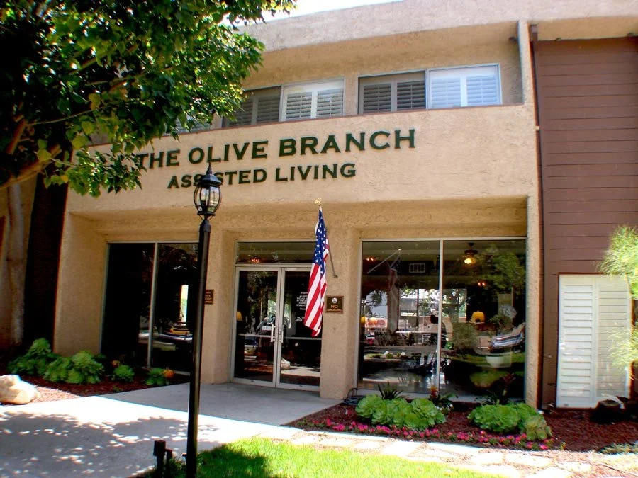 Exterior view of The Olive Branch Assisted Living in Northridge featuring a beige facade, American flag, landscaped entryway, and a welcoming, residential-style entrance.