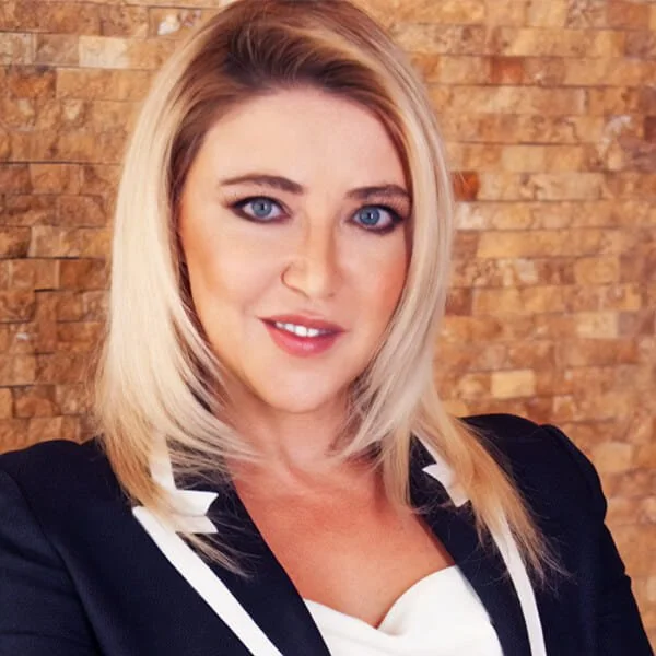 Professional woman with blond hair in a dark blazer, posing against a stone wall.
