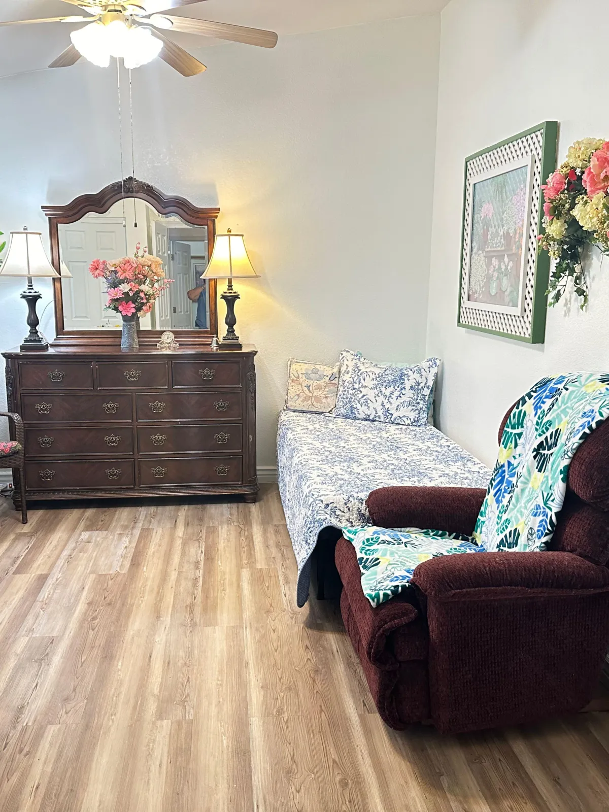 A cozy resident room at Active Assisted Care in Scottsdale, featuring a floral-covered twin bed, elegant wooden dresser with lamps, a cushioned recliner with a colorful throw, and homey wall decor.