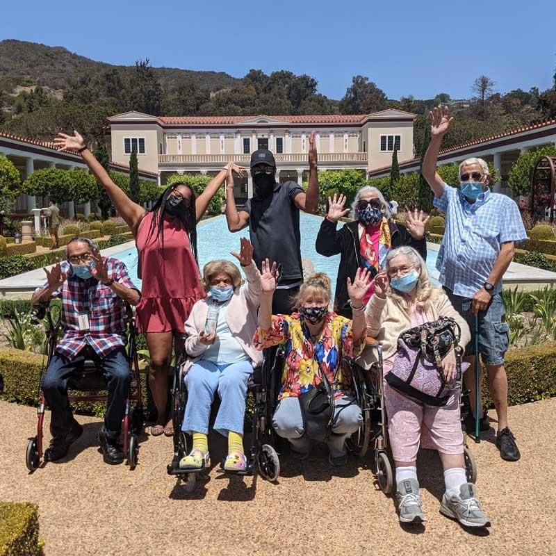 A group of seniors and caregivers enjoying an outdoor outing at a scenic location with a grand estate and mountains in the background