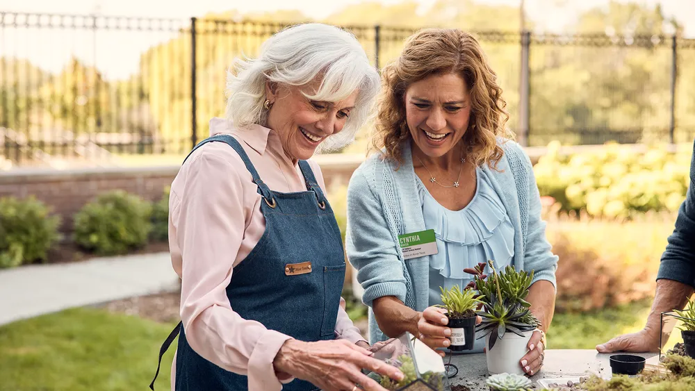 Two women, one of whom is an Atria employee, smile and garden outdoors at a senior living community.