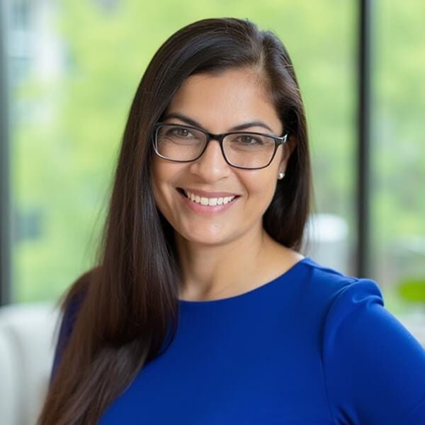Smiling woman with long dark hair and glasses in a blue dress in a bright office.