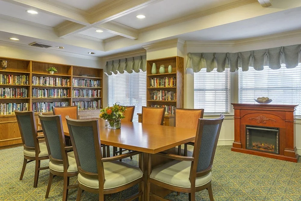 A cozy senior living library with a large wooden table, comfortable chairs, built-in bookshelves filled with books, and a fireplace, creating a warm and inviting space for residents at Brookdale North Scottsdale