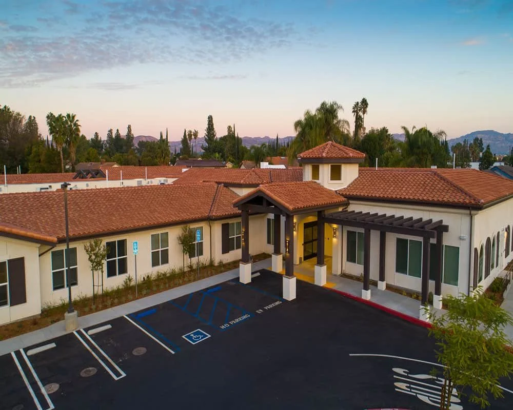 Front view of The Preserve at Woodland Hills assisted living and memory care community in Los Angeles, featuring Spanish-style architecture, red tile roofing, accessible parking, and surrounding palm trees at sunset.