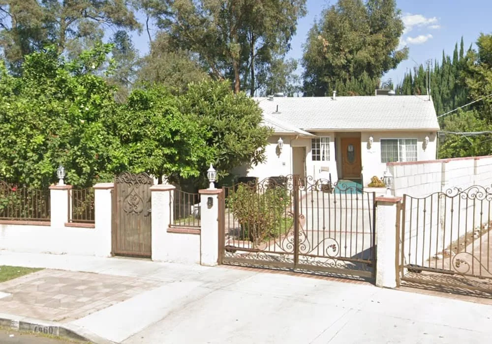 Front exterior of A Compassion Valley assisted living home in Van Nuys, a one-story white house behind a white wall and wrought-iron gate with dense green trees around it.