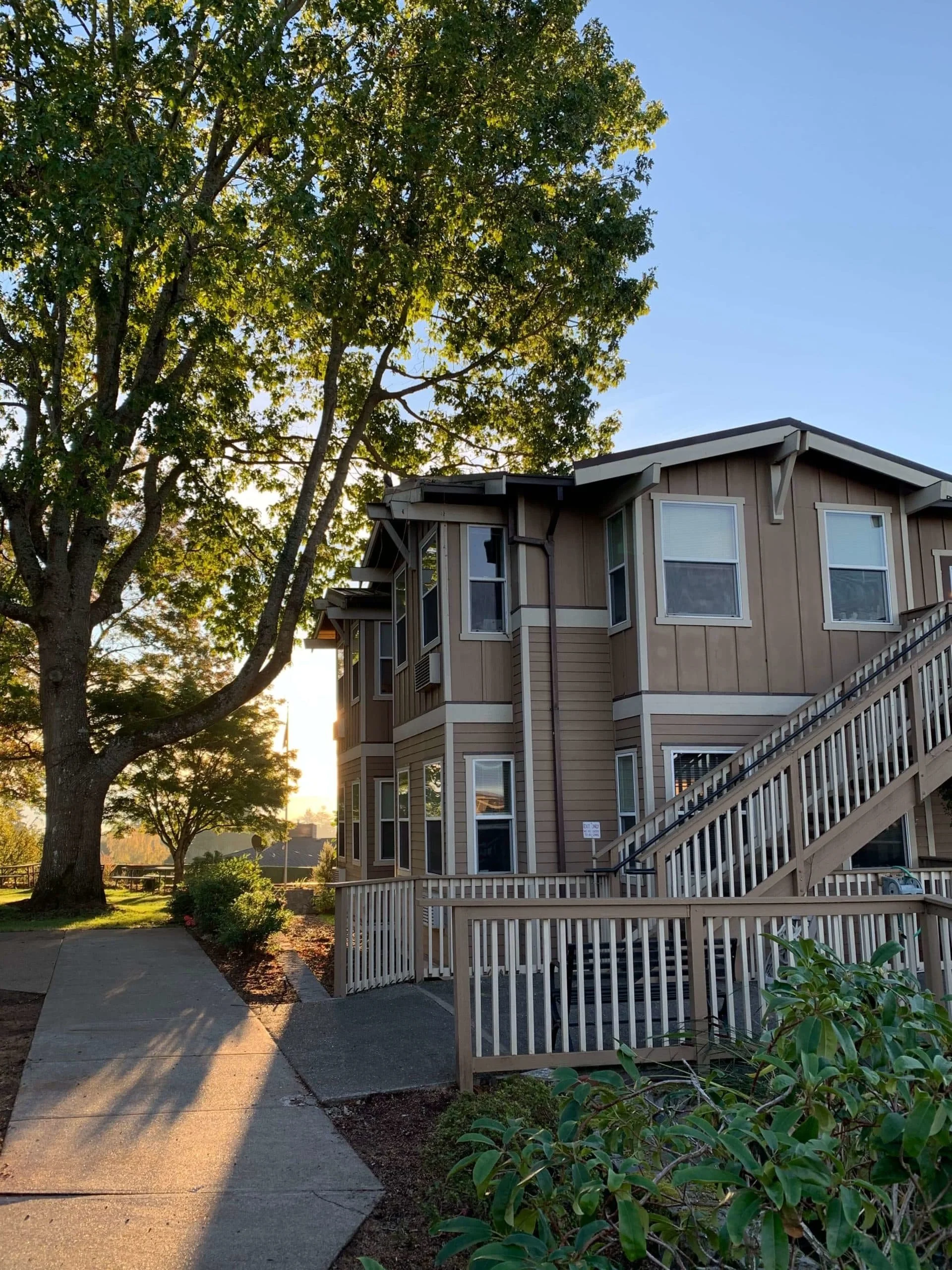Modern two-story memory care facility surrounded by trees and evening sunlight, featuring wheelchair-accessible ramps and a peaceful outdoor setting