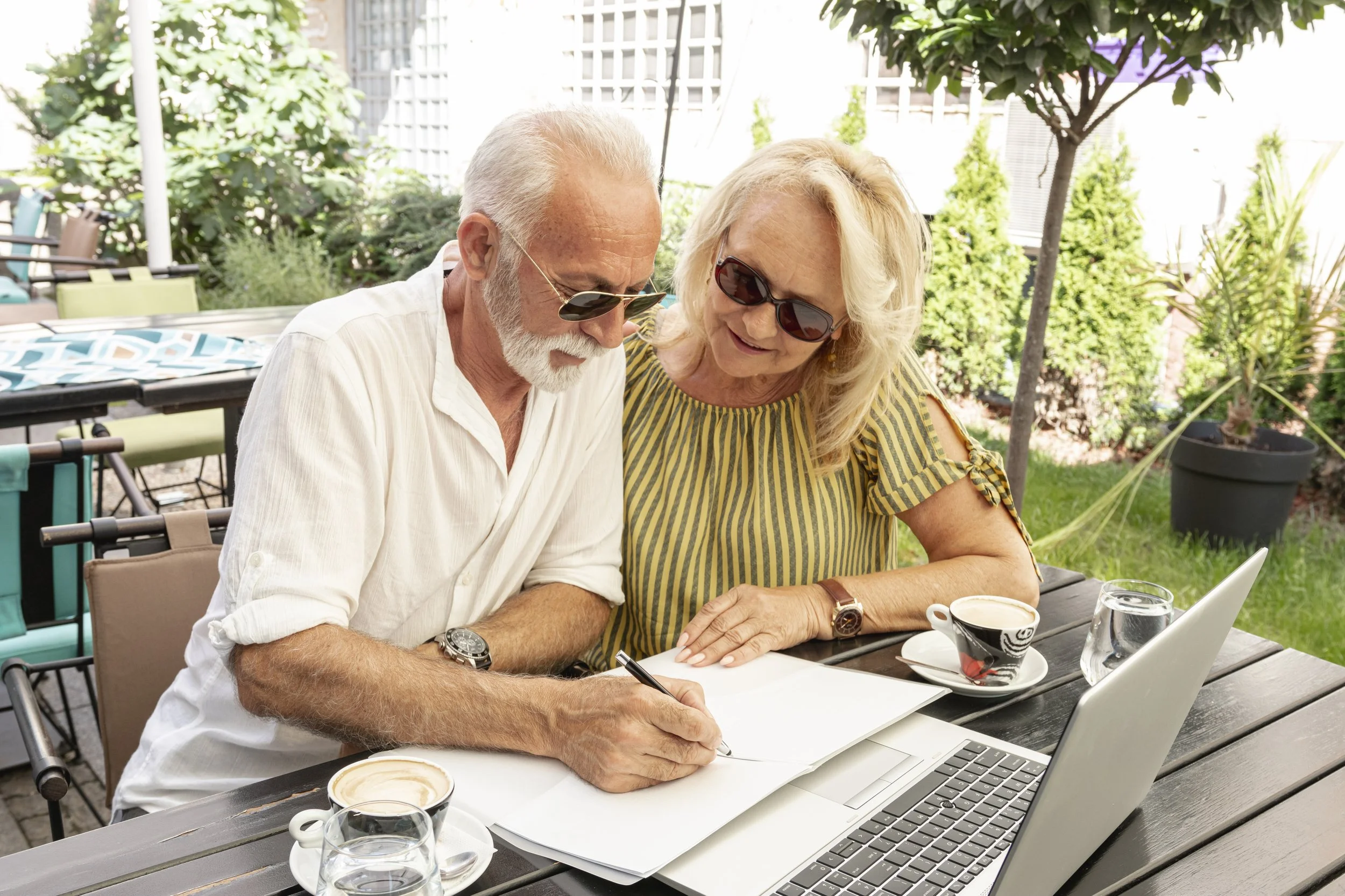 Senior couple sitting outdoors at Savant Burbank West, reviewing notes and enjoying coffee together, surrounded by greenery and sunshine in a relaxed courtyard setting.