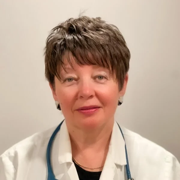 Experienced female clinician in white coat with stethoscope, neutral studio background.