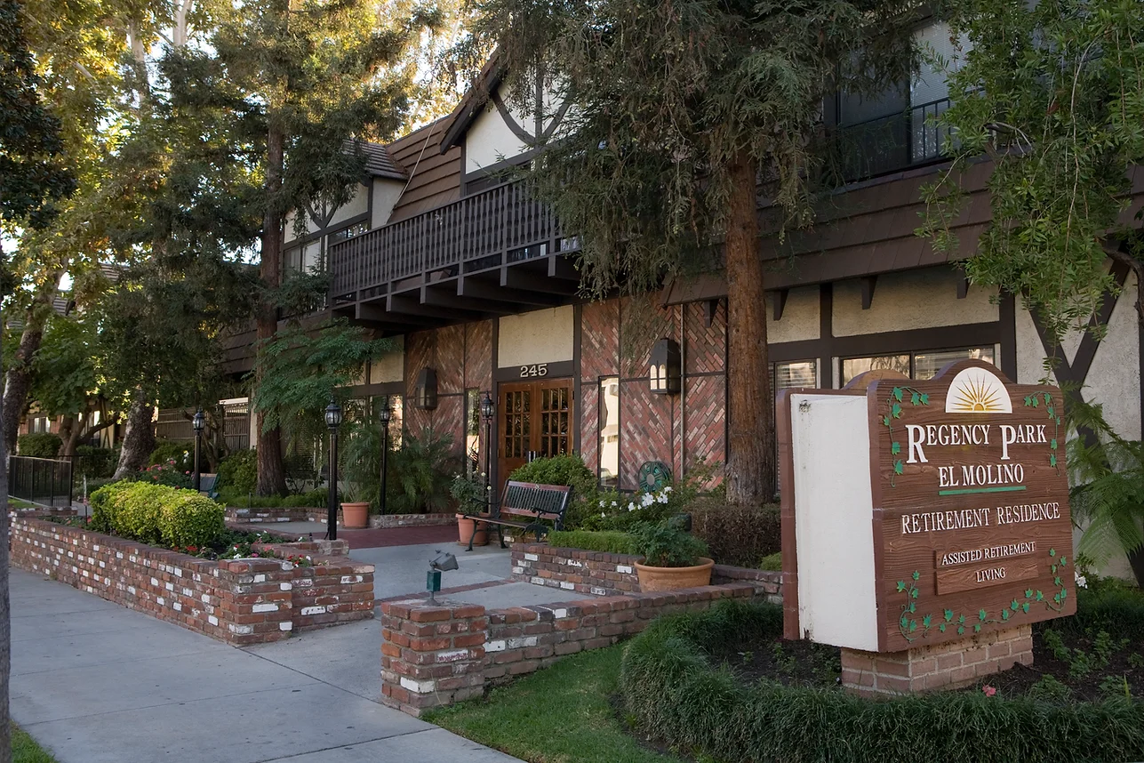 Front exterior of Regency Park El Molino retirement residence, featuring Tudor-style architecture, brick walls, a lush landscaped entrance with benches and greenery, creating a warm and inviting atmosphere for assisted living residents.