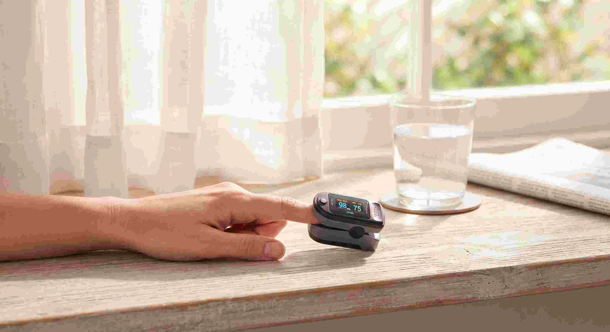 Fingertip pulse oximeter resting on a sunlit windowsill beside a glass of water