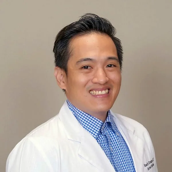 Male physician in white coat and blue checkered shirt smiling in front of a neutral wall.