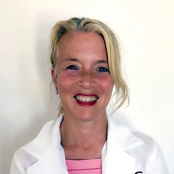 Smiling female doctor in white coat and pink top against a light background.