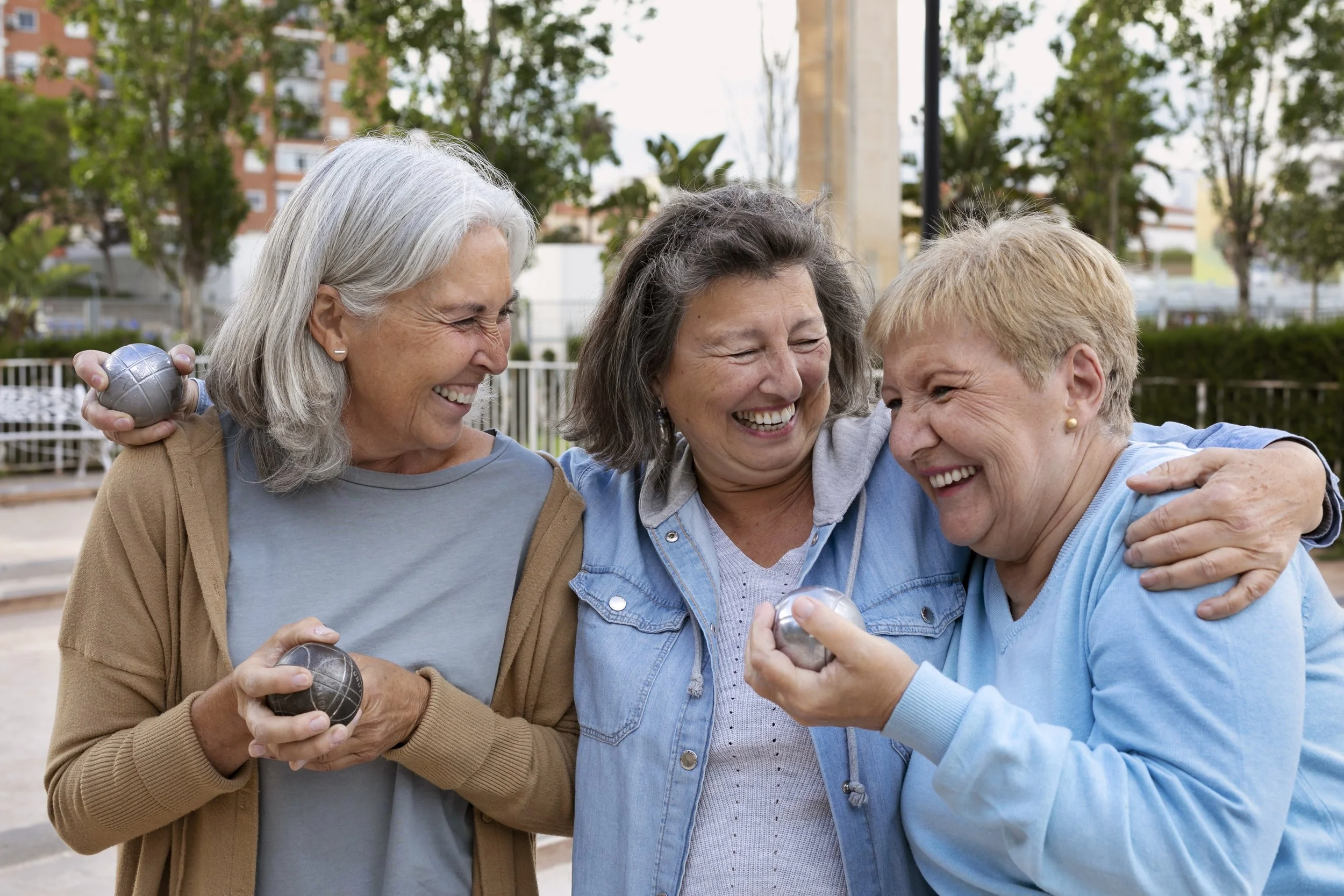 Three senior women laughing and embracing outdoors while playing a game of pétanque at Active Care Homes in Scottsdale, enjoying social time and fresh air together.