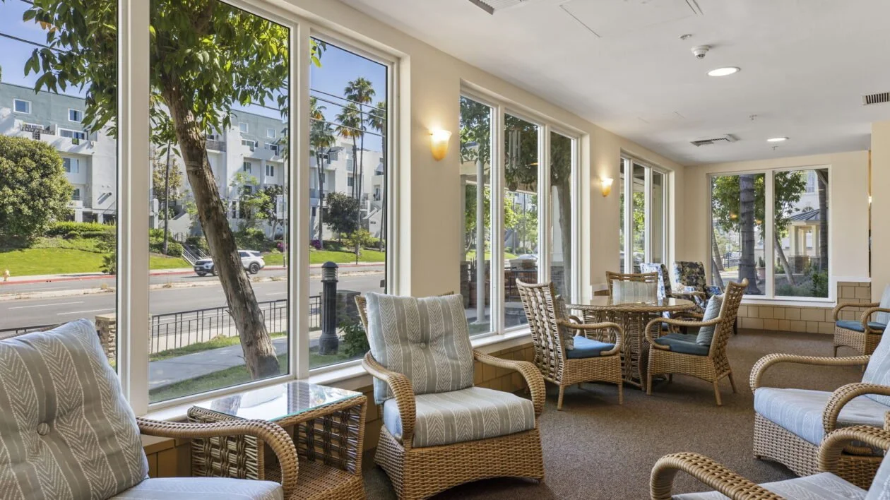 Bright and inviting sunroom at Ivy Park at Woodland Hills with wicker chairs and large windows overlooking a green, tree-lined street, offering residents a peaceful space to relax and socialize