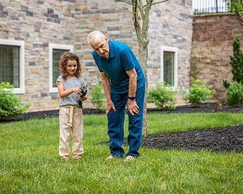 An elderly man in a blue outfit enjoying time outdoors with a young girl holding a small black puppy, standing on a lush green lawn in front of a stone building