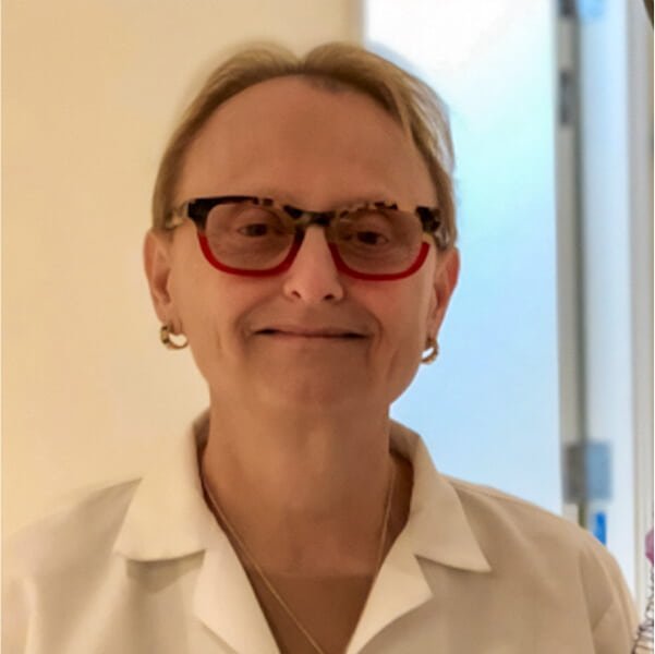 Smiling older doctor in glasses and white coat standing in a bright hallway.