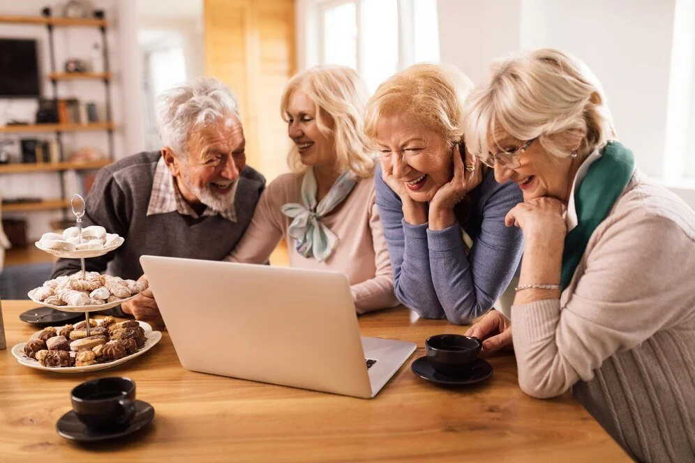 Group of smiling seniors gathered around a laptop, enjoying coffee and desserts, illustrating the social connectivity and joyful community life at MPTF Los Angeles.