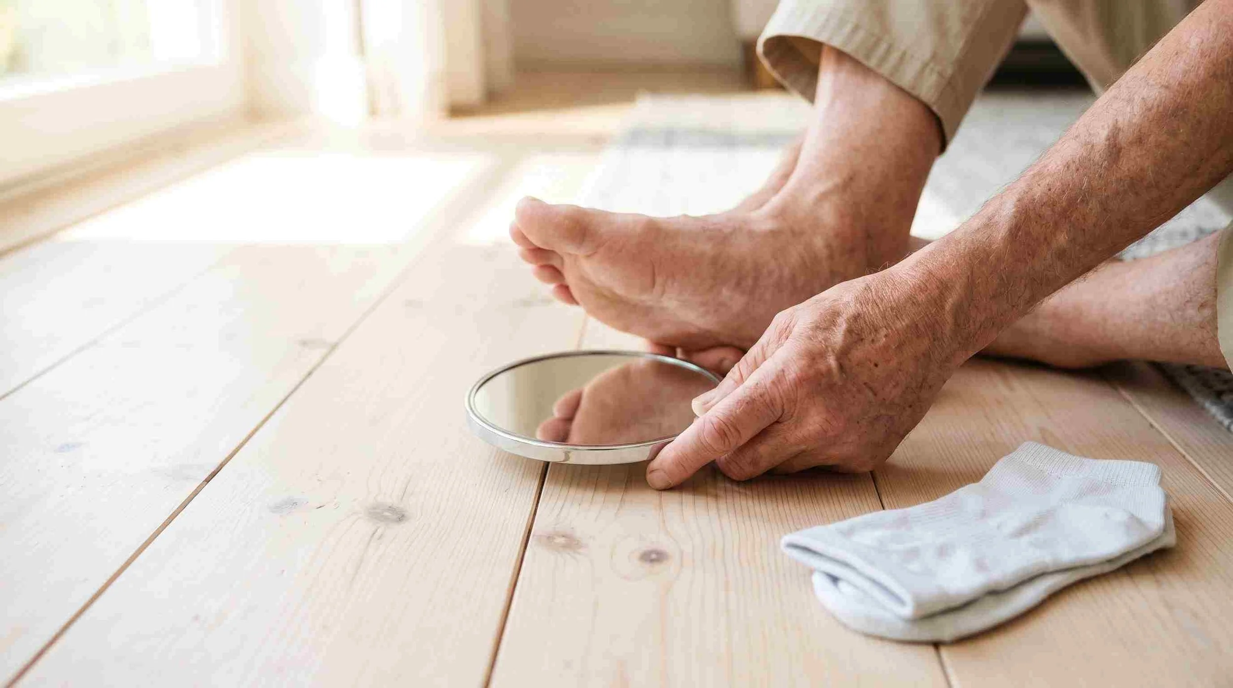 Older man using a small hand mirror on the floor to check the sole of his bare foot in morning light