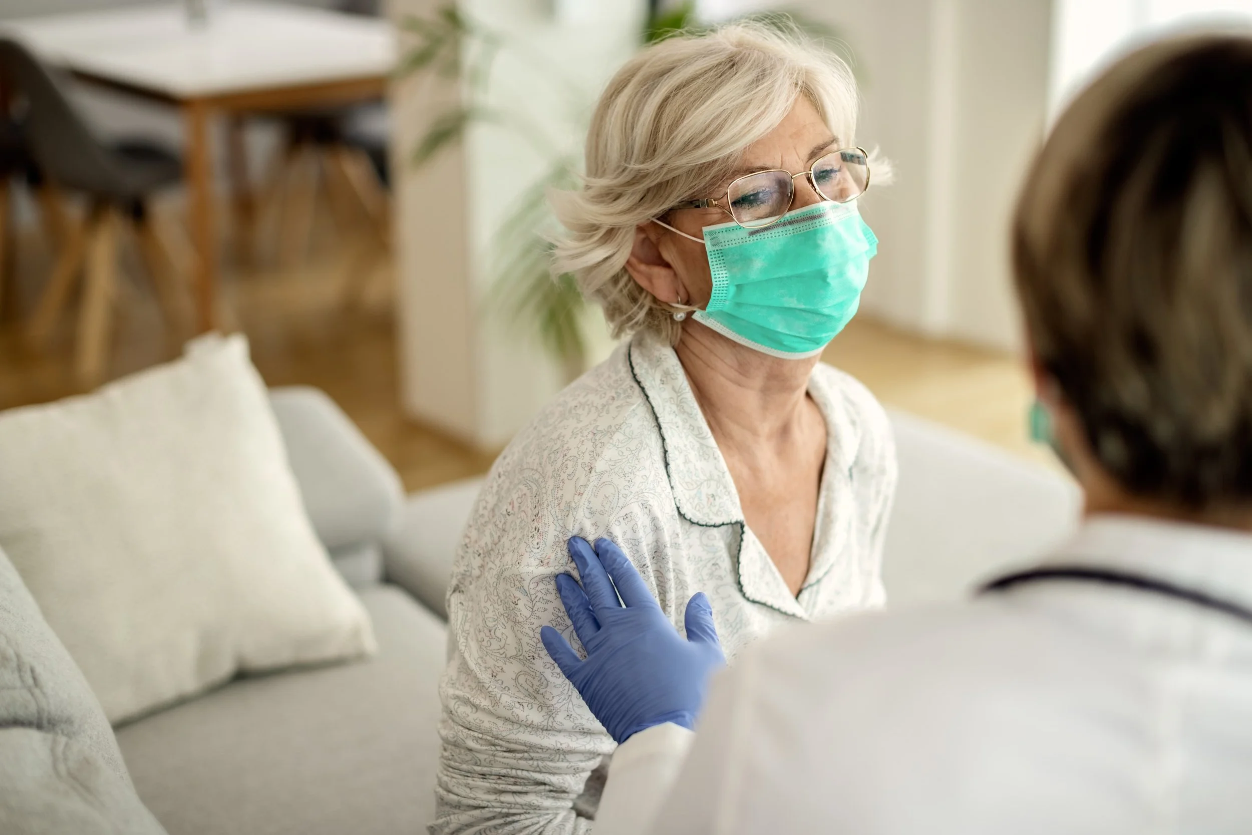 Elderly woman wearing a protective face mask while receiving in-home medical consultation from a healthcare provider.