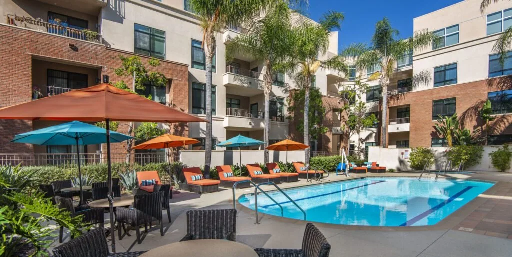 Outdoor pool and lounge area at The Village at Northridge, featuring shaded seating, palm trees, and a modern resort-style building in a senior living community.