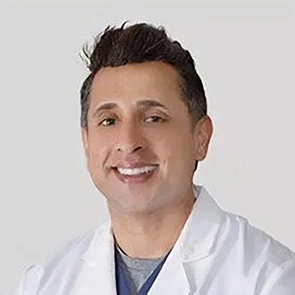 Male doctor in white coat smiling against a light background.