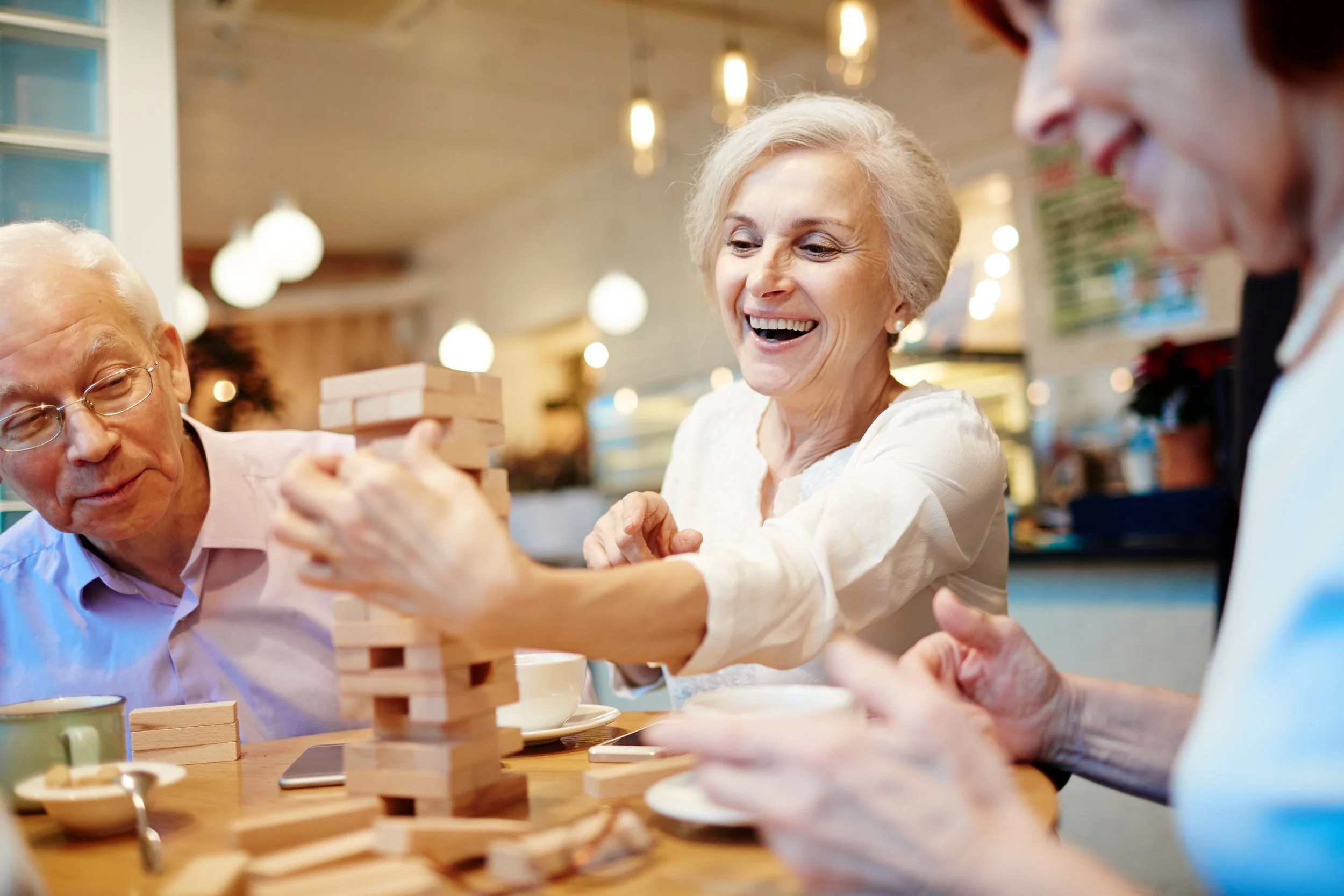 Smiling elderly residents enjoying a game of Jenga together in a cozy, well-lit common area at A Happy Place Adult Care Home, fostering joy and connection.