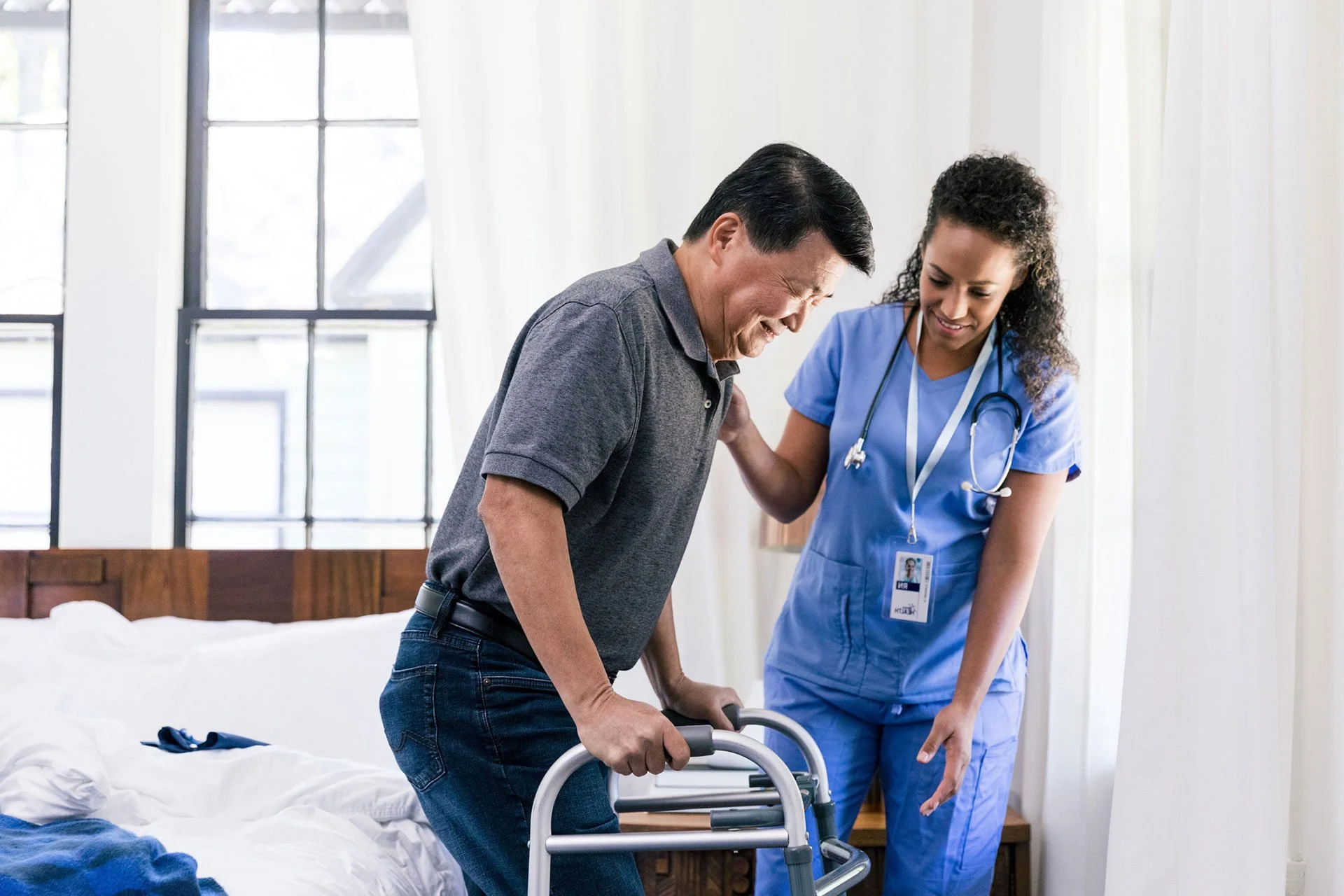 A compassionate healthcare professional in blue scrubs assists an elderly man using a walker during a rehabilitation session, providing support and encouragement in a bright and comfortable setting.
