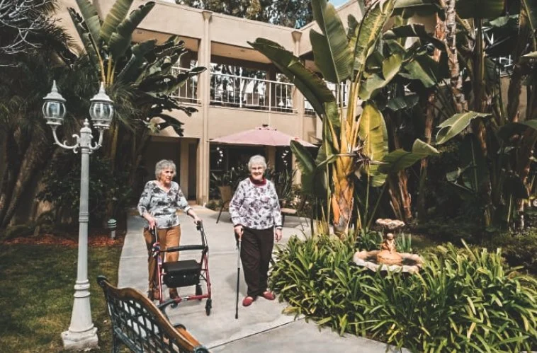 Two senior residents enjoying a walk in the lush outdoor garden of Park Balboa, surrounded by tropical greenery and a peaceful fountain. The well-maintained courtyard provides a serene and relaxing environment for socializing and outdoor activities.