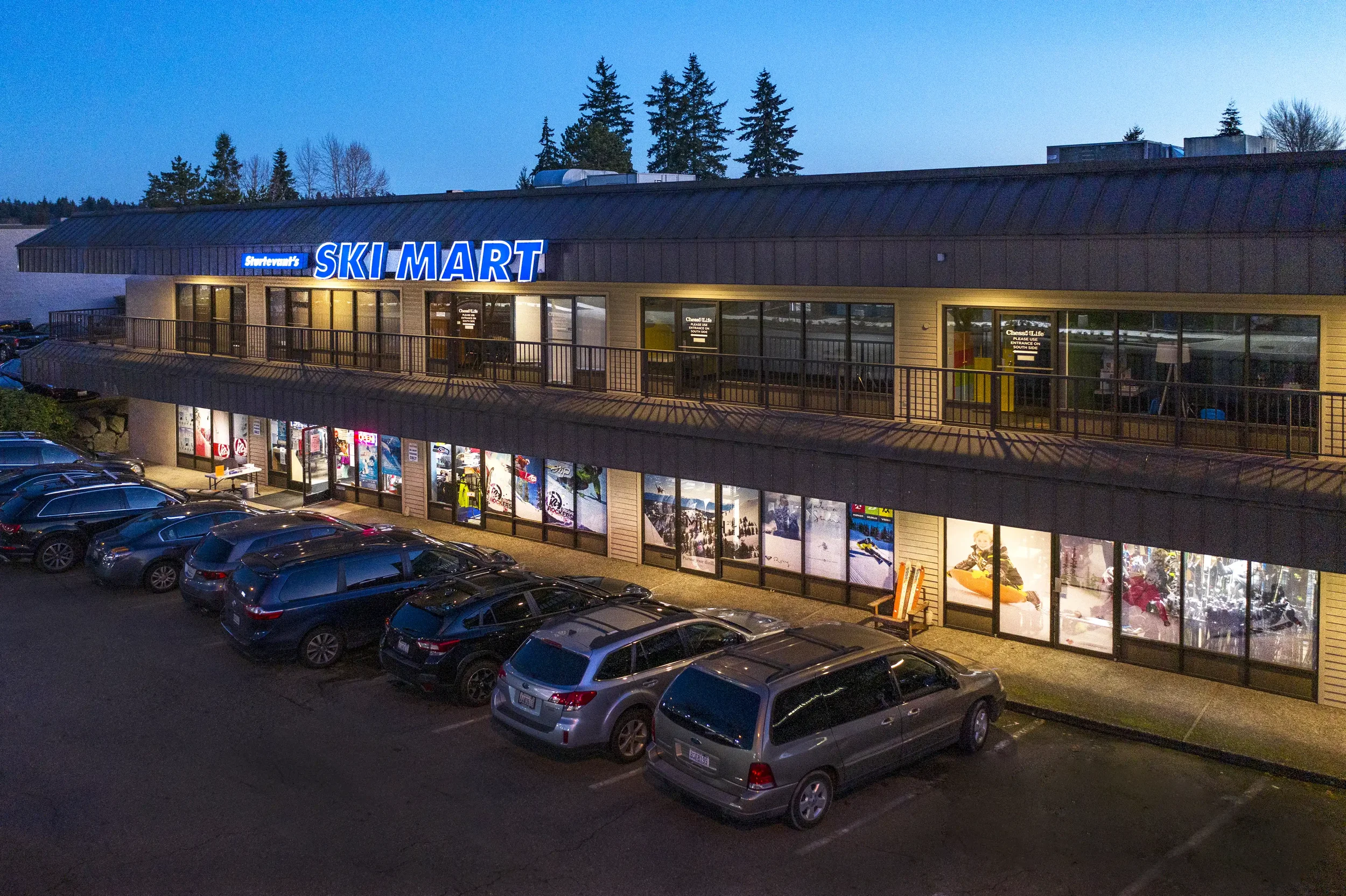 A two-story building with a prominent blue sign reading 'Ski Mart' and an upper walkway with large windows, parked cars in front, and a background of trees at dusk.