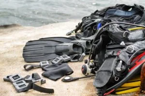 Collection of scuba diving gear on a sandy beach, including fins, masks, snorkels, weight belts, and wetsuits.