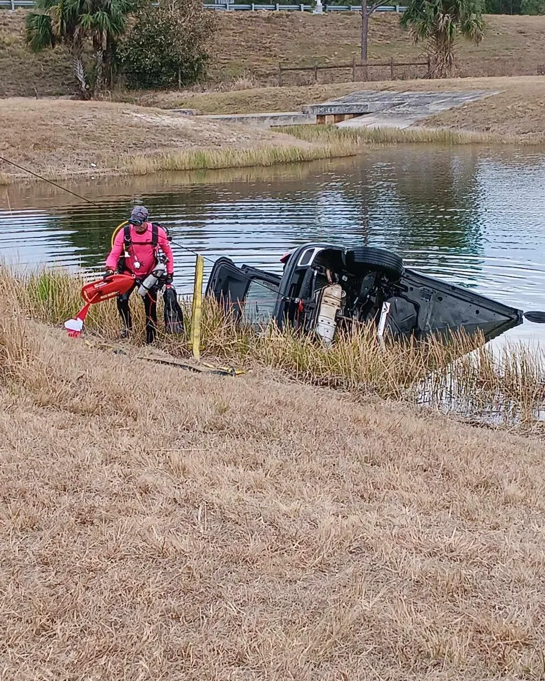 A person in pink gear and helmet standing on grassy bank near a partially submerged black SUV in a body of water, possibly after an accident.