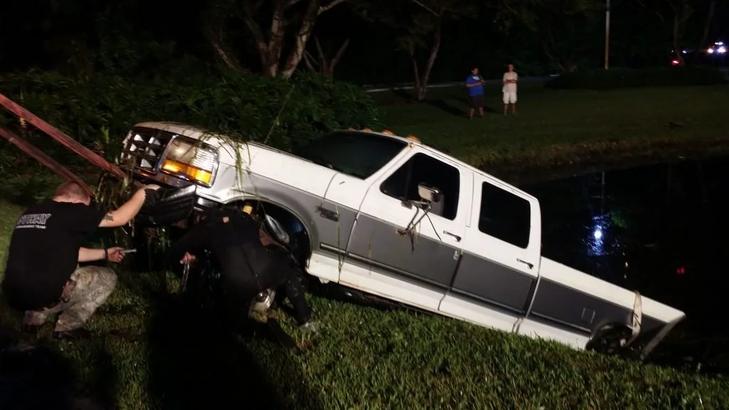 A white truck has fallen into a body of water with emergency responders examining the scene during nighttime.