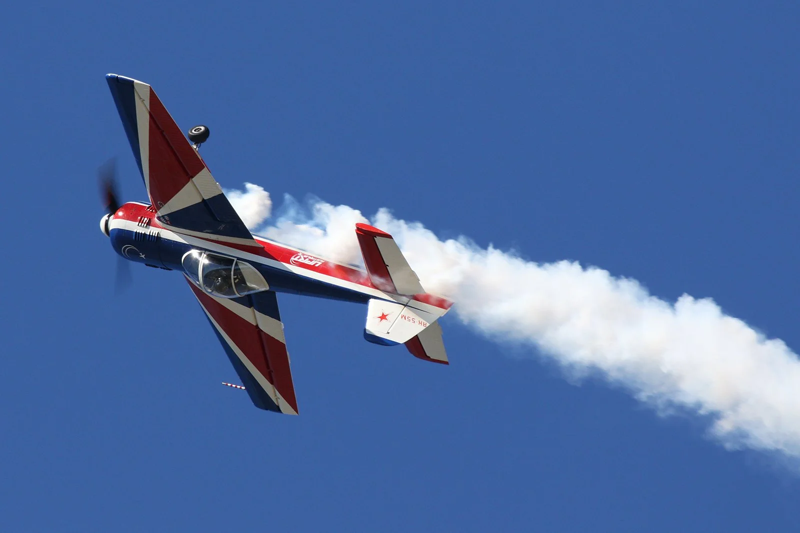 A small propeller-driven aircraft with a polished silver body painted with red, white, and blue stripes, on a runway with grass and trees in the background, with the propeller spinning quickly.