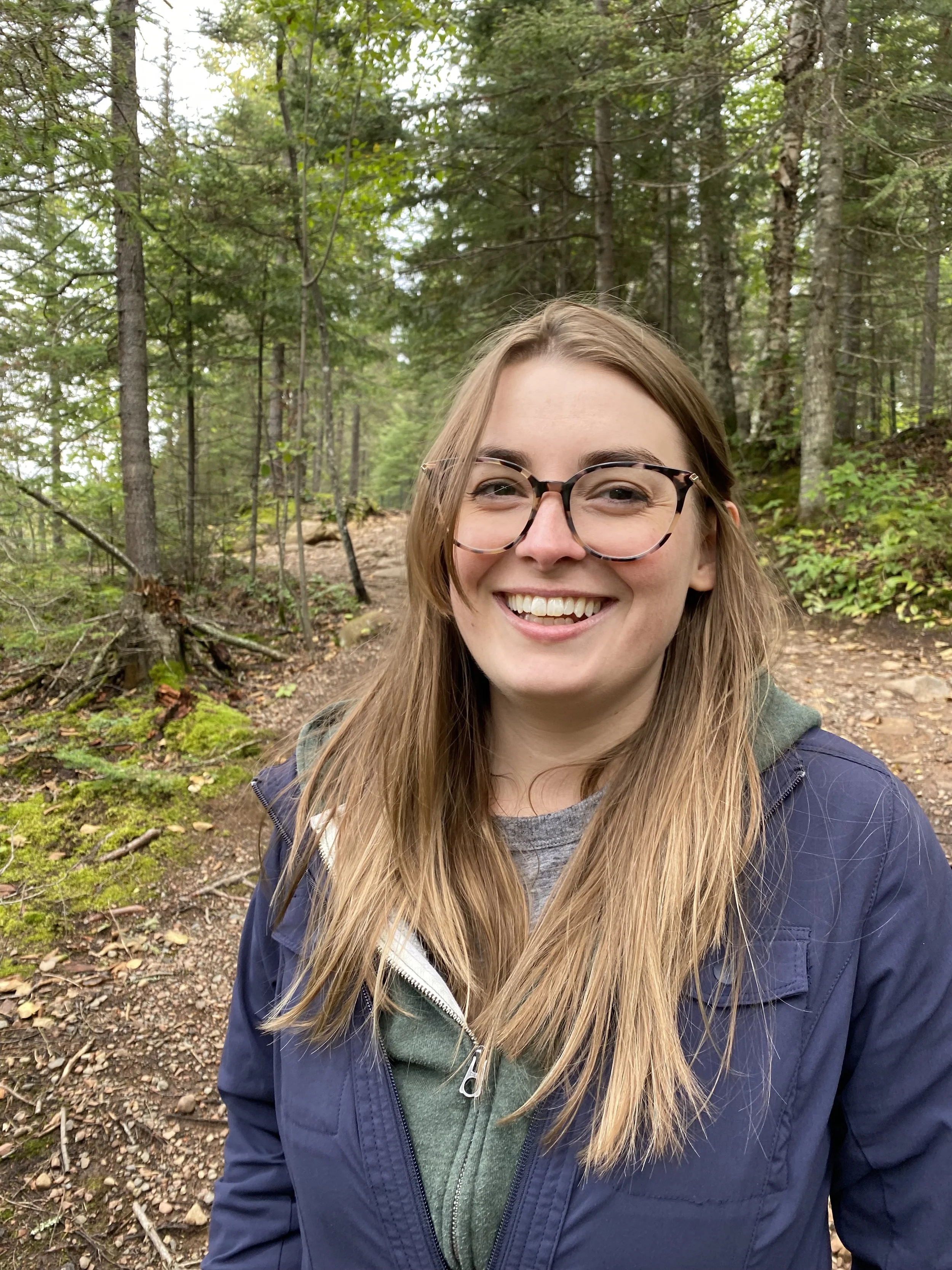 Woman with long brown hair and glasses smiling at the camera. Background is a hiking trail surrounded by trees.