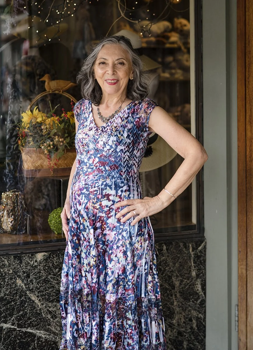 Mary Ellen Grills, owner of Peppergrass & Tulip of 40 years, wearing a colorful patterned dress, smiling and standing in front of a store window display with flowers and decorative items.