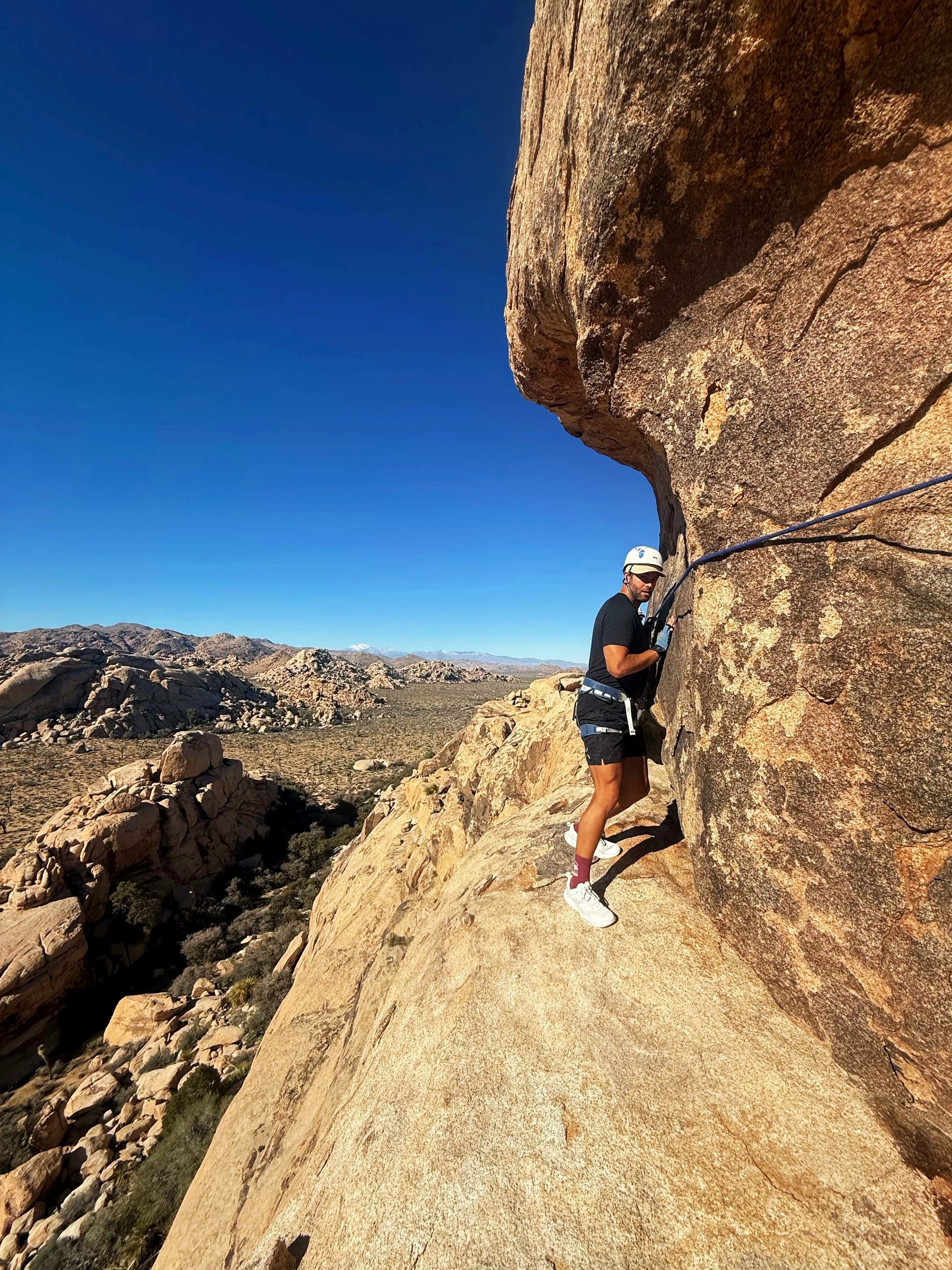 A person climbing a rock face outdoors with a harness and helmet in a desert landscape under a clear blue sky.