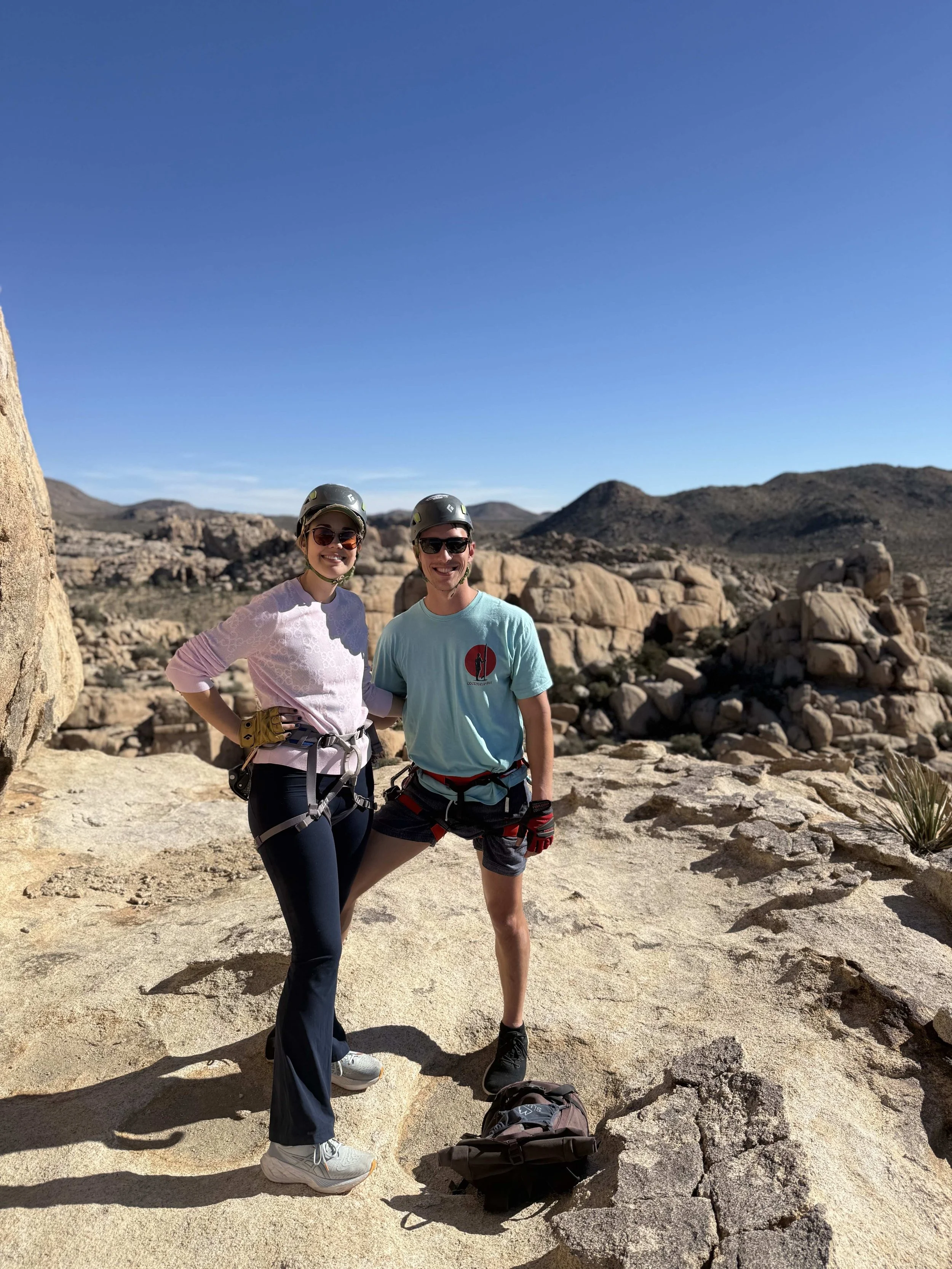 Two climbers wearing helmets and harnesses standing on a rocky desert landscape, smiling at the camera with a backpack on the ground in front of them.