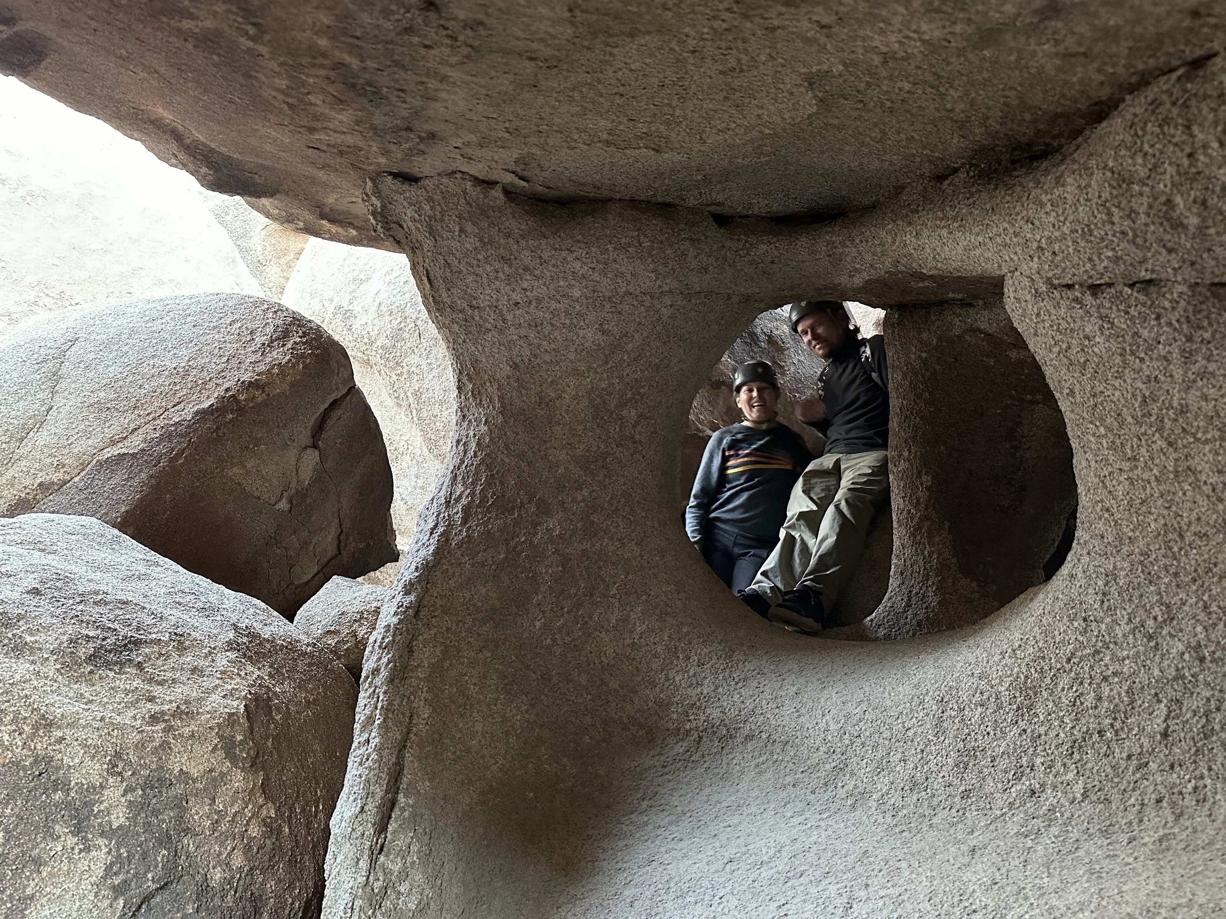 Two people with helmets smiling inside a circular rock formation in a natural canyon.