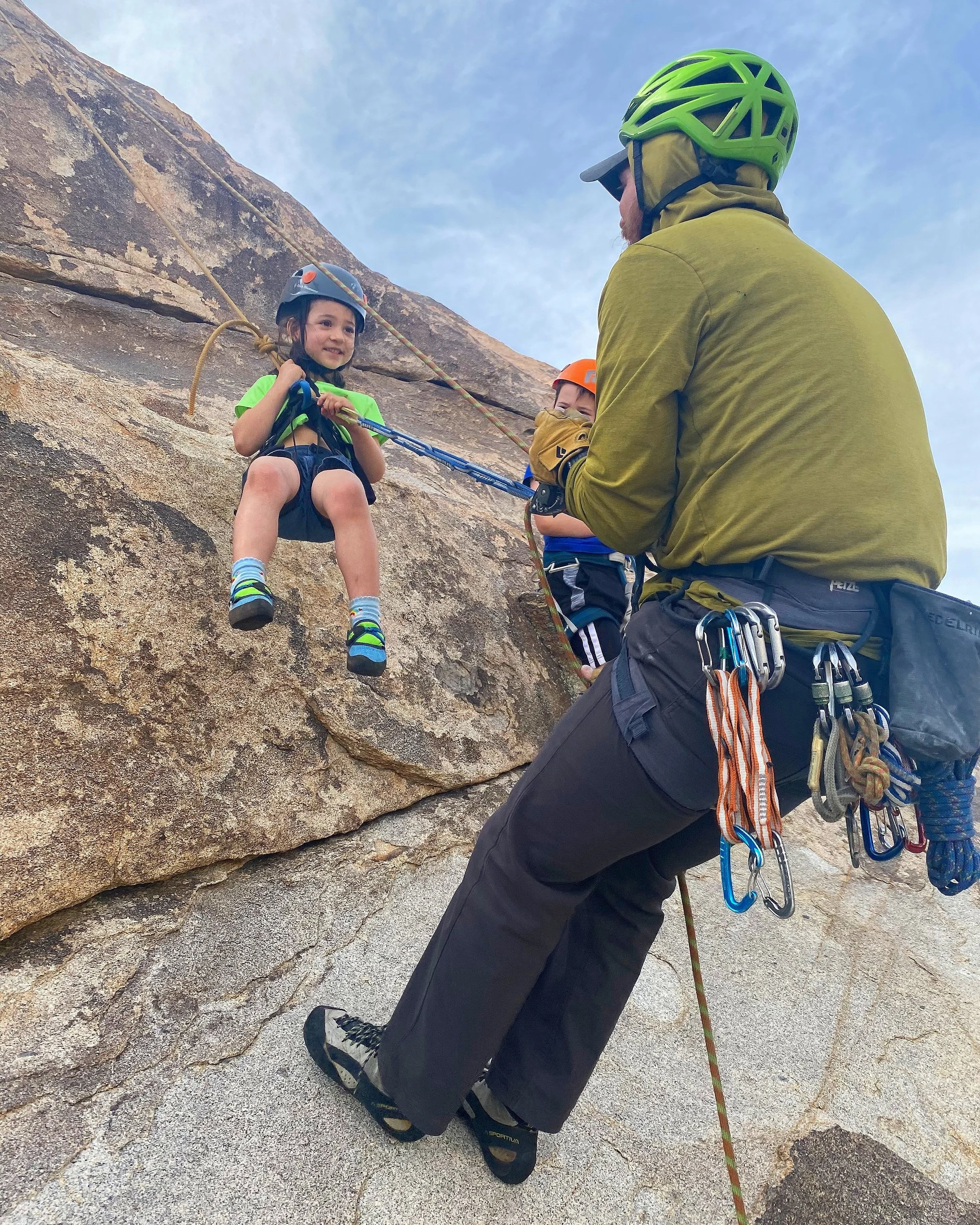 A young girl smiling while rock climbing with a helmet and harness, assisted by an instructor on a rock face outdoors.