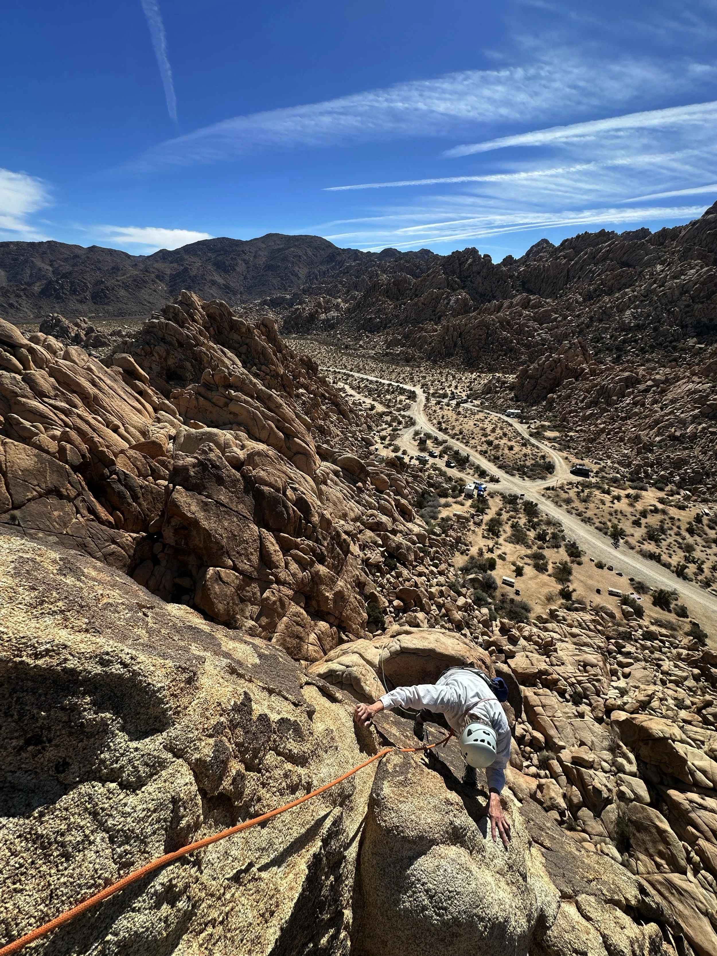 A person in a white shirt and helmet rock climbing on a steep rocky desert mountain with a dirt road and parked cars below, under a bright blue sky with wispy clouds.