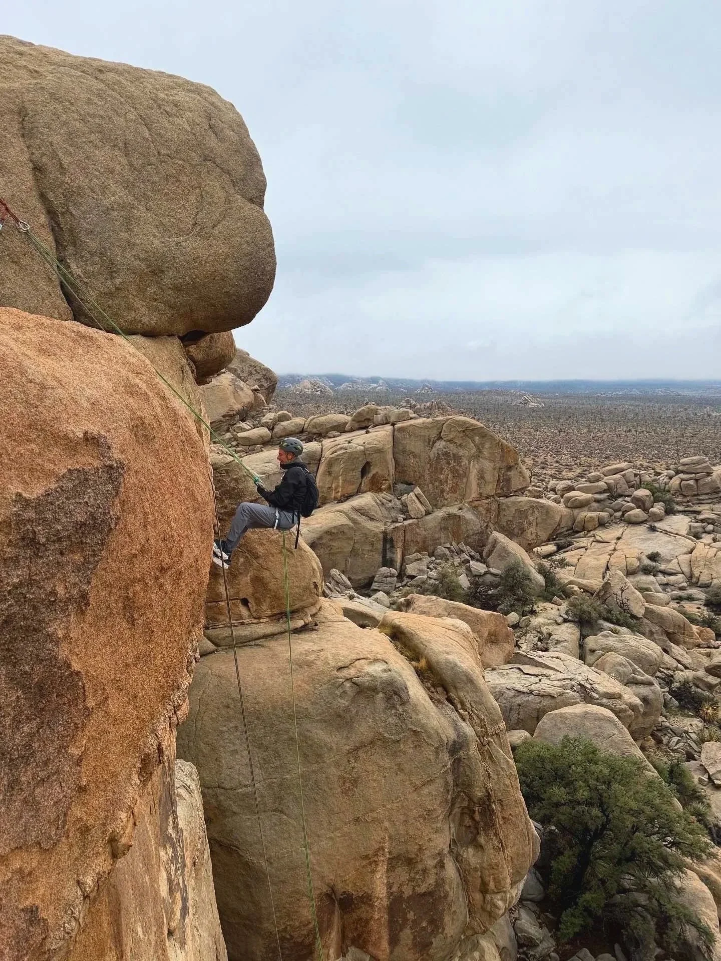 A person wearing a helmet and harness climbing a large rock formation in a desert landscape with scattered boulders and sparse vegetation under a cloudy sky.