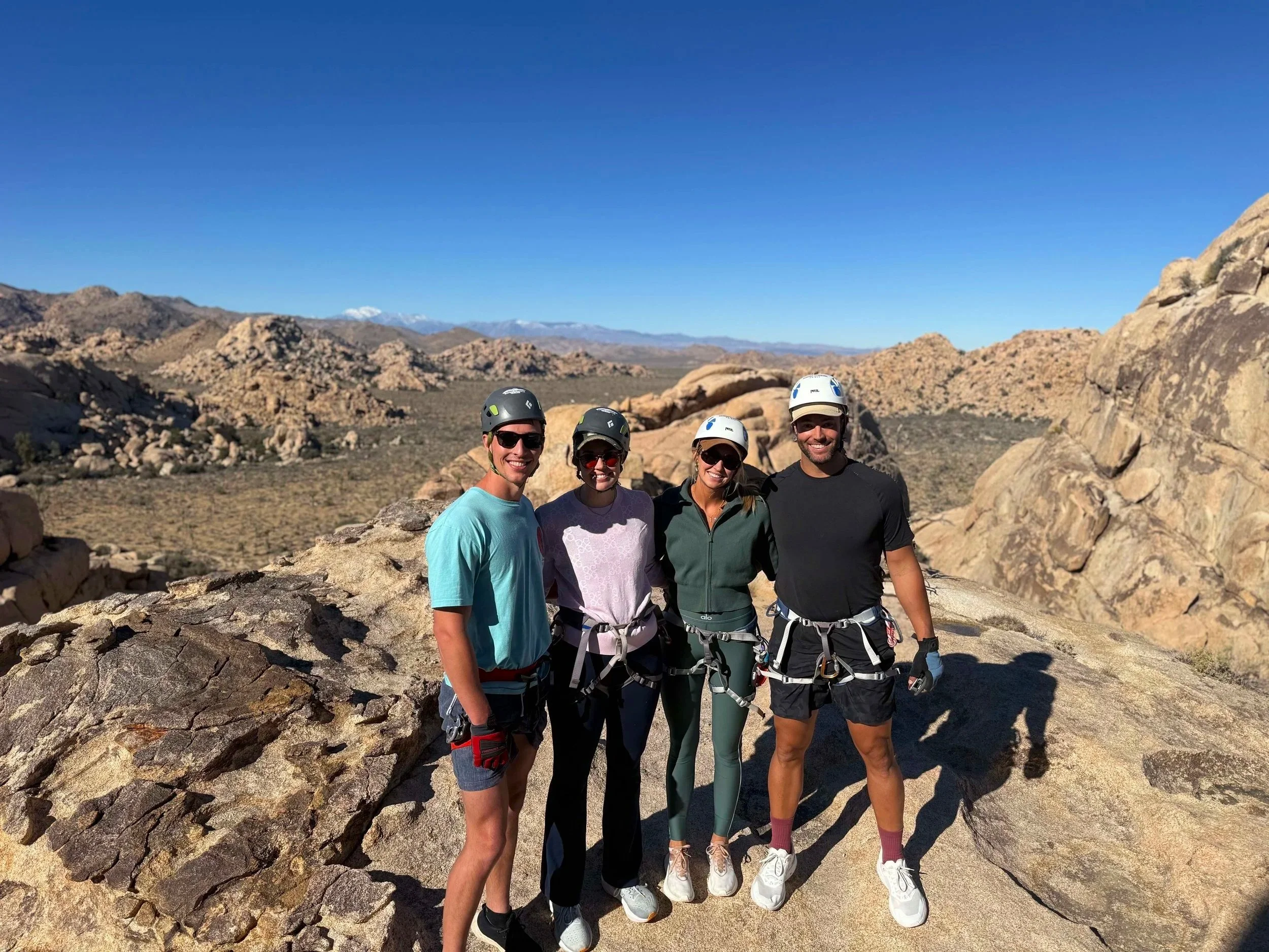 Four people standing on a large rock in a desert landscape with mountains in the background, wearing helmets and climbing harnesses.