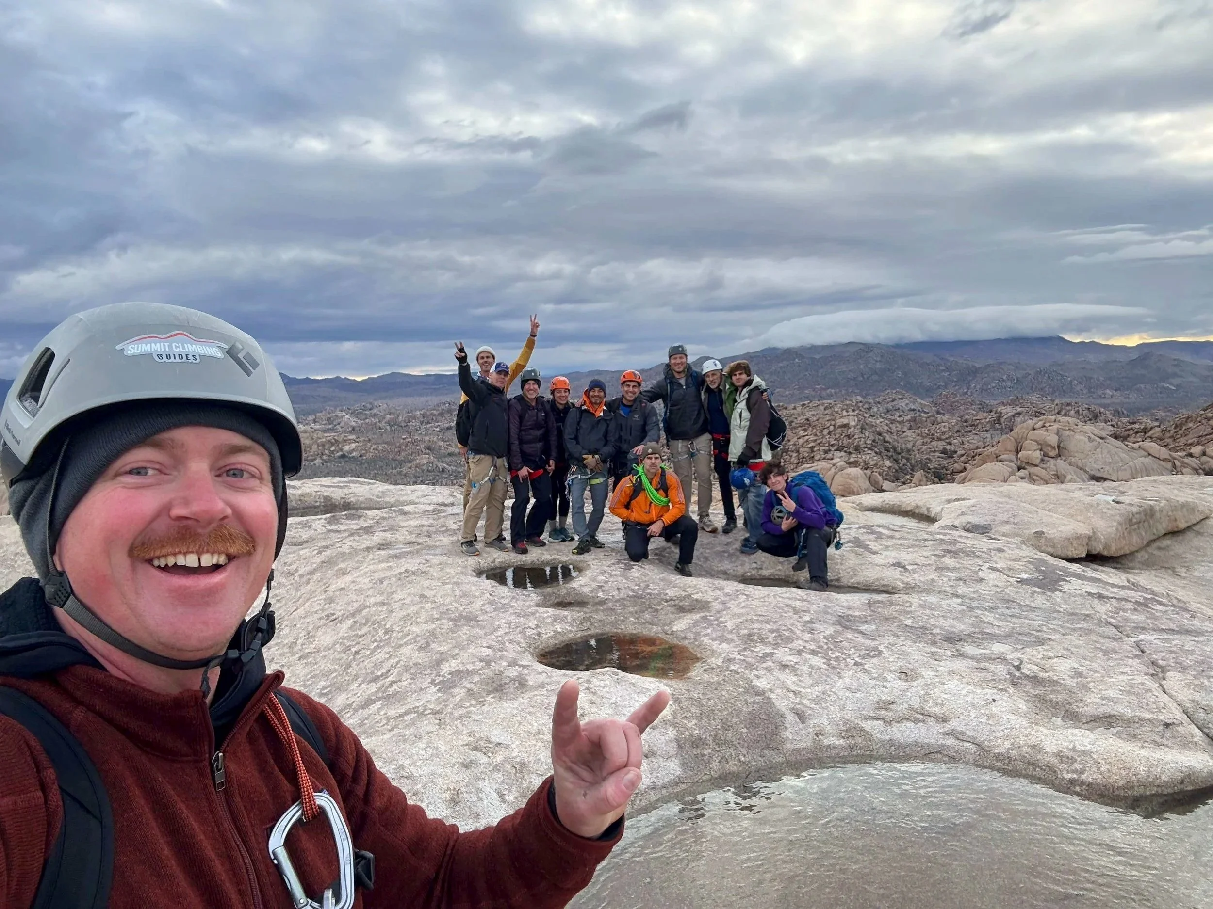 A group of people on a mountain summit, one person in the foreground wearing a helmet and smiling, pointing at a small water pool on the rocky surface, with mountains and cloudy sky in the background.