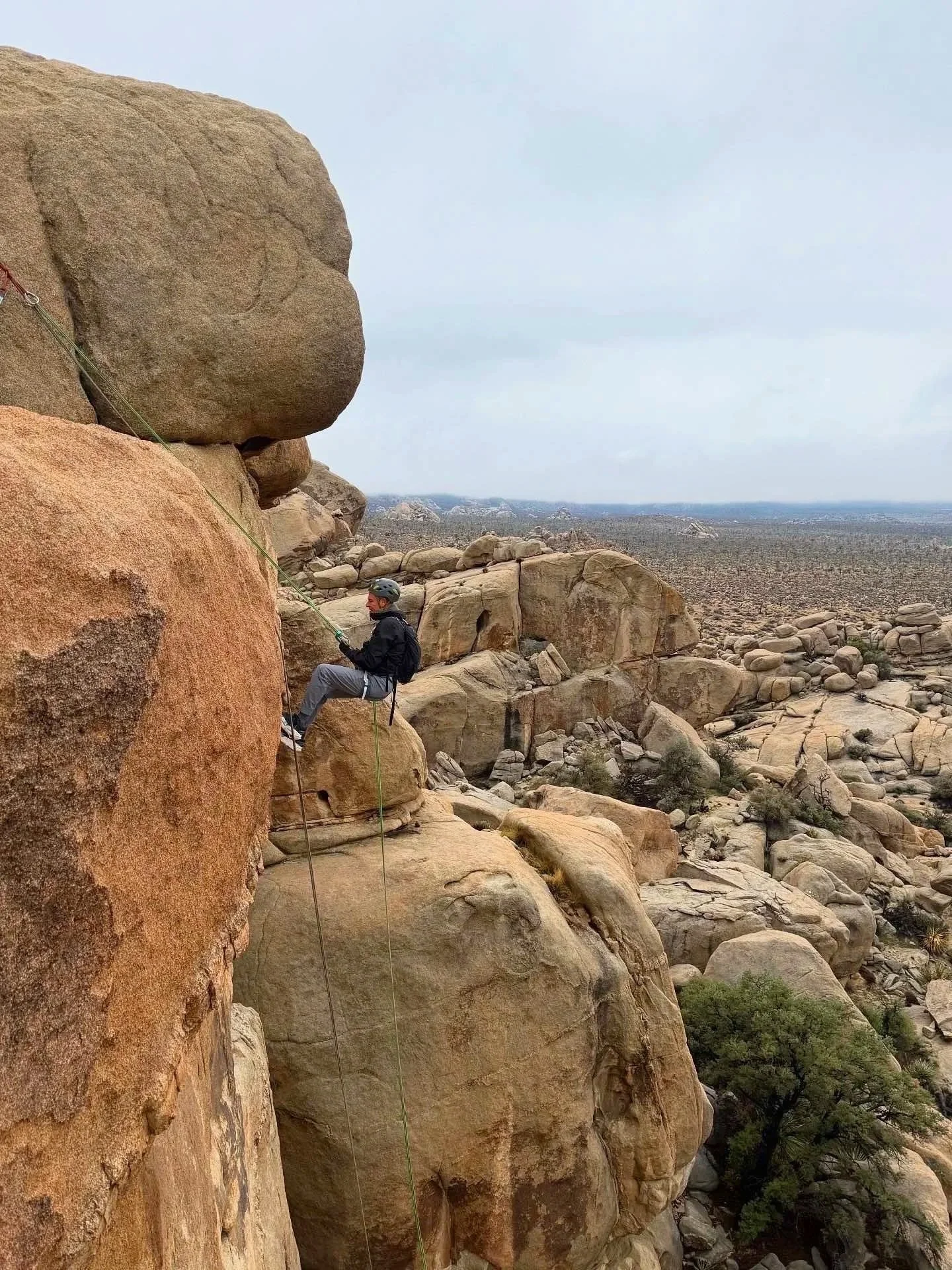 A person rock climbing or rappelling on a large boulder in a desert landscape with many rocks and sparse vegetation under a cloudy sky.